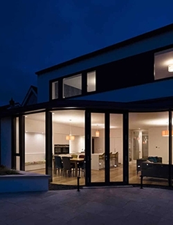 Evening view of a modern residential home extension in South Dublin with expansive glass doors and windows illuminated from within, showcasing an inviting domestic interior, under a twilight sky