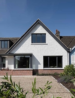 White-rendered gable-fronted house in Terenure, Dublin with contrasting black window frames, featuring a bay window on the ground floor, set against a brick driveway and surrounded by green shrubbery. Simple residential architecture.