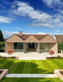 Contemporary brick residence with a symmetrical architectural design, large glass doors, and a central chimney, complemented by a manicured lawn and neatly arranged shrubs, under a clear blue Dublin sky